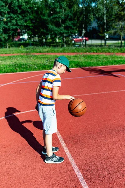 Beyaz tenli küçük basketbolcu açık havada kırmızı kortta antrenman yapıyor.