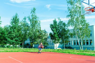 Telsiai, Litvanya 08: 12 2020 Çekici Kafkasyalı küçük basketbolcu açık havada kırmızı kortta antrenman yapıyor.