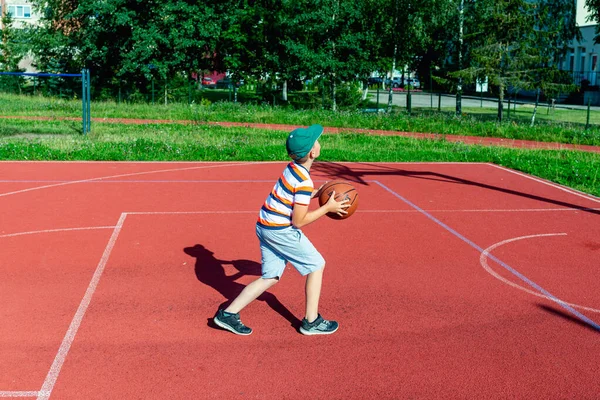 Telsiai, Litvanya 08: 12 2020 Çekici Kafkasyalı küçük basketbolcu açık havada kırmızı kortta antrenman yapıyor.