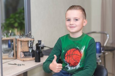 Bir çocuk saç tıraşından sonra oturup kuaförde başparmağını gösteriyor. Mutlu çocuk iyi bir hareket gösteriyor..