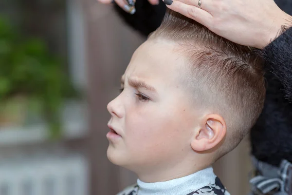 Hairdresser makes a stylish hairstyle. The woman is standing and making haircut for blonde boy. Close up side view.