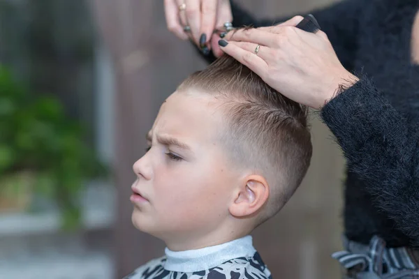 Hairdresser makes a stylish hairstyle. The woman is standing and making haircut for blonde boy. Side view image.