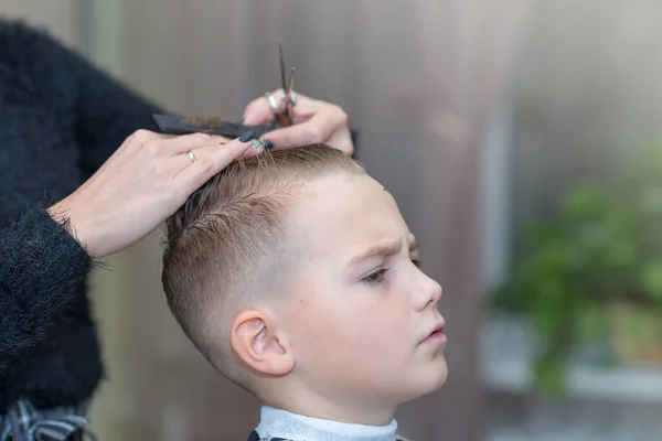 Hairdresser makes a stylish hairstyle. The woman is standing and making haircut for blonde boy. Side view image.