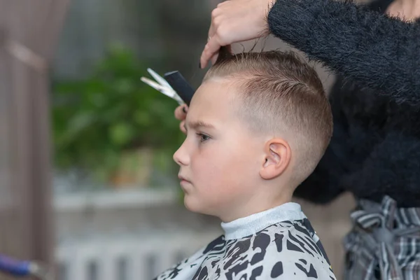 Hairdresser makes a stylish hairstyle. The woman is standing and making haircut for blonde boy. Side view image.