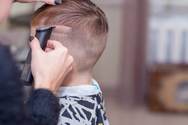 Hairdresser makes a stylish hairstyle. The woman is standing and making haircut for blonde boy. Close up of hands.