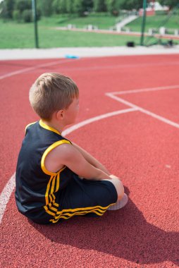 Yandan bakıldığında üzgün ilkokul çocuğu basketbol sahasında oturuyor.