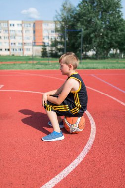 Basketbolun üzerinde oturan üzgün ilkokul çocuğunun yan görüntüsü