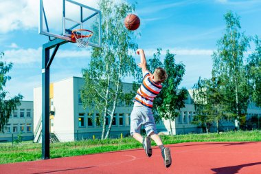Çekici beyaz küçük basketbolcu topu potaya atmak için atlıyor. Açık havada kırmızı kortta.