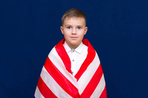 快乐的年轻的高加索裔美国男孩儿用美国国旗包裹着自己 用蓝色背景的相机看着自己 — 图库照片