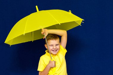 Mavi stüdyo arka planında şemsiyesi olan beyaz, sarışın, çekici bir öğrenci. Çocuk her türlü somurtmayı gösteriyor..