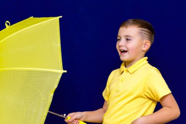 Side view Attractive caucasian blond schoolboy with umbrella on blue studio background.Boy shows all kinds of grimaces. clipart