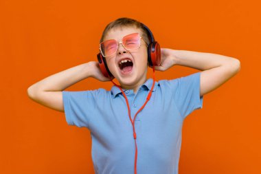 Kırmızı gözlüklü çekici çocuk turuncu stüdyo arka planında kırmızı kulaklıklı müzik dinliyor.