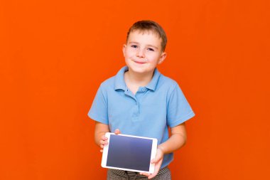 Avrupalı öğrencinin portresi turuncu stüdyo arka planında tablet ve mavi tişört gösteriyor. Neşeli bir şekilde bir şeyler düşünüyor ve boş tablet ekranında bir şeyler gösteriyor. Turuncu zemin. Anlaşıldı, afiş..