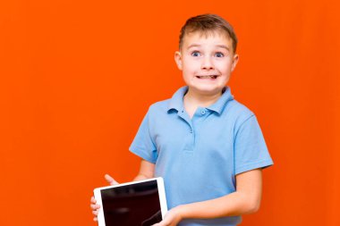 Avrupalı öğrencinin portresi turuncu stüdyo arka planında tablet ve mavi tişört gösteriyor. Neşeli bir şekilde bir şeyler düşünüyor ve boş tablet ekranında bir şeyler gösteriyor. Turuncu zemin. Anlaşıldı, afiş..