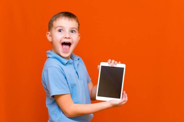 Şaşırmış okul çocuğunun portresi turuncu stüdyo arka planında tablet ve mavi tişört gösteriyor. Sevinçle bir şeyler düşünüyor ve boş tablet ekranında bir şeyler gösteriyor. Turuncu arka plan..