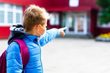 Kapalı çekimde koruyucu maskeli çekici okul çocuğu dışarıda dikilirken okuldaki heyecanlı çocuk parmakla işaret ederek okula geri dönüyor. Salgın grip ve koronavirüs.