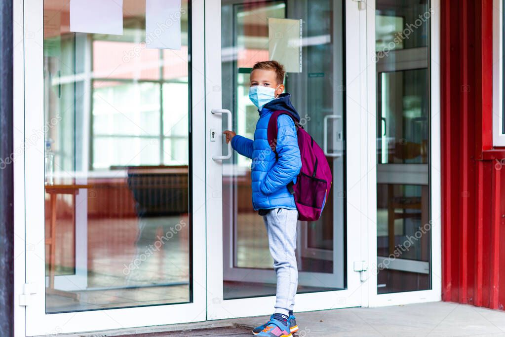 Full length side view schoolboy wearing protective mask is trying to open the school door. Behind the backpack Schoolboy look at camera