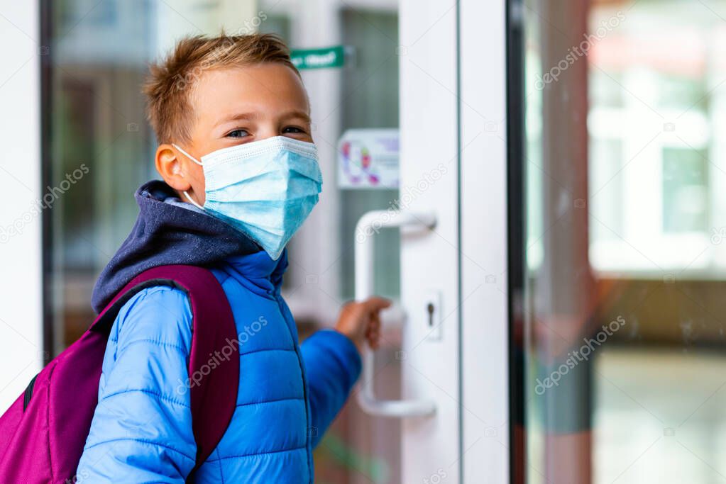 Young schoolboy wearing protective mask is trying to open the school door. Behind the backpack Schoolboy look at camera