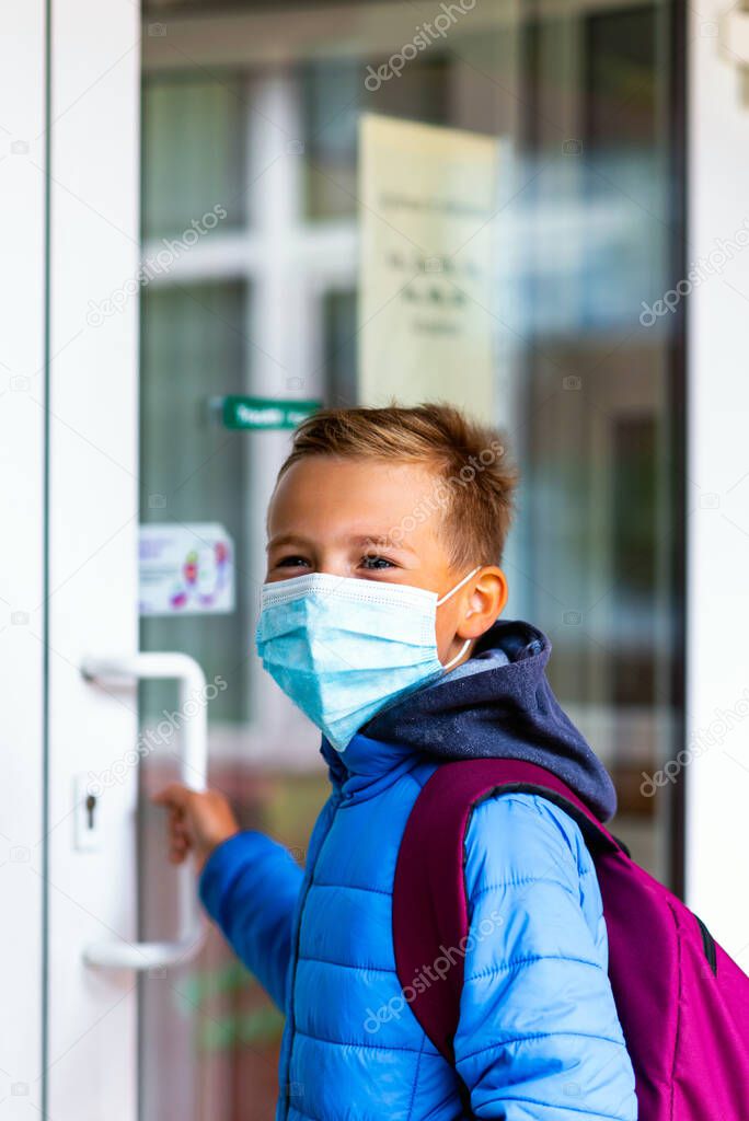 side view young schoolboy wearing protective mask is trying to open the school door. Behind the backpack Schoolboy look aside