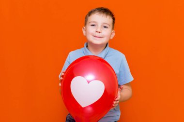 Sevgililer Günü 'nü kapat. Kafkasyalı neşeli çocuk elinde büyük kırmızı bir balon tutuyor. Balonda beyaz kalpli bir çocuk. Turuncu stüdyo arka planı..
