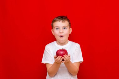 Avrupalı çocuk okul çocuğu, elinde kırmızı bir elma elması tutuyor. Vitaminler, meyveler, sağlıklı yiyecekler. Parlak kırmızı arka planda mutlu, güzel bir çocuğun önsözü.