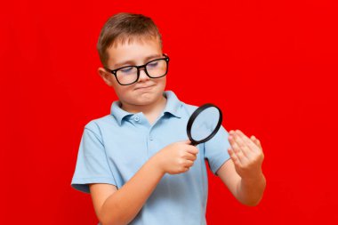 Gözlüklü ve büyüteçle avucunun içine bakan güzel Avrupalı genç bir çocuk. Kırmızı stüdyo duvarı..