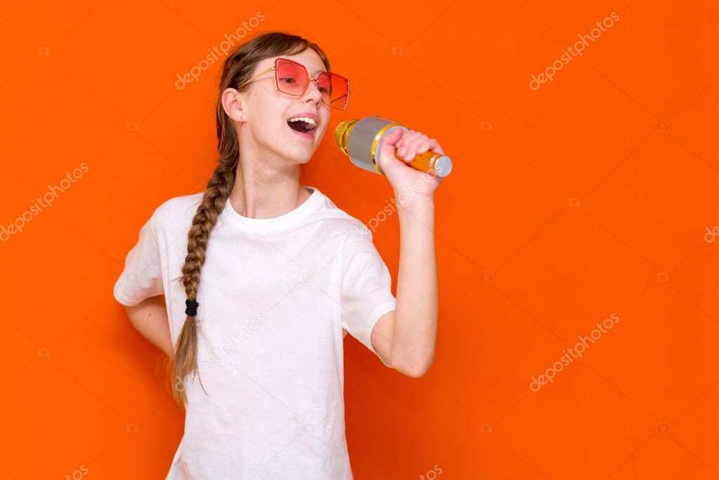 Happy young beautiful girl in red sunglasses singing with microphone on orange studio background