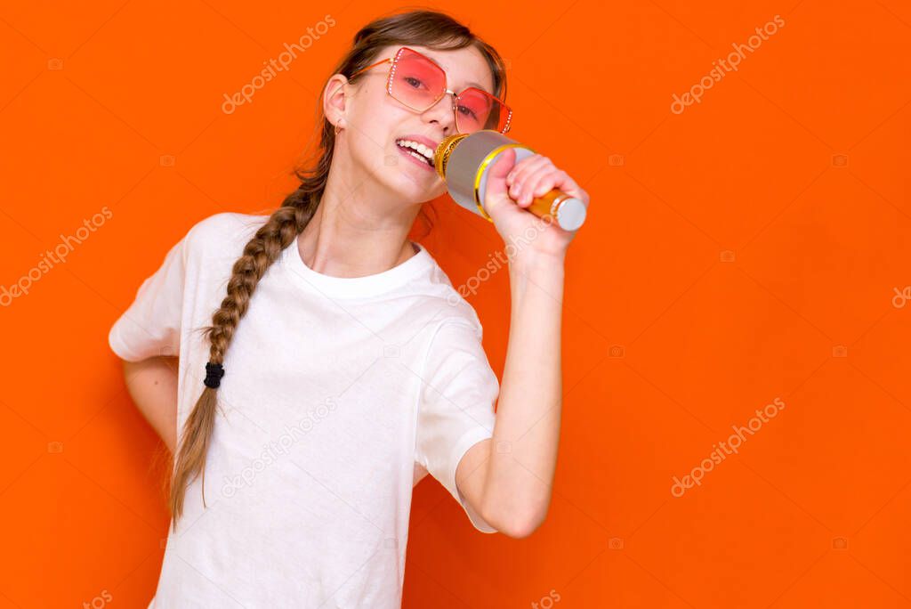 Happy young beautiful girl in sunglasses singing with microphone on orange studio background