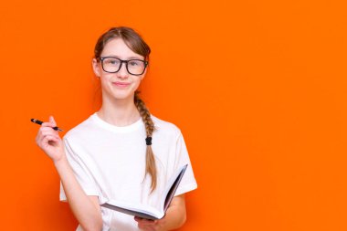 Gözlüklü, beyaz tişörtlü, kalemli, gözlüklü genç bir kıza bakarken heyecanlanıyorum. Bir şeyler düşün ve turuncu bir stüdyonun arka planındaki bir deftere bak. Mesajın, afişin, ilanın için yer aç..