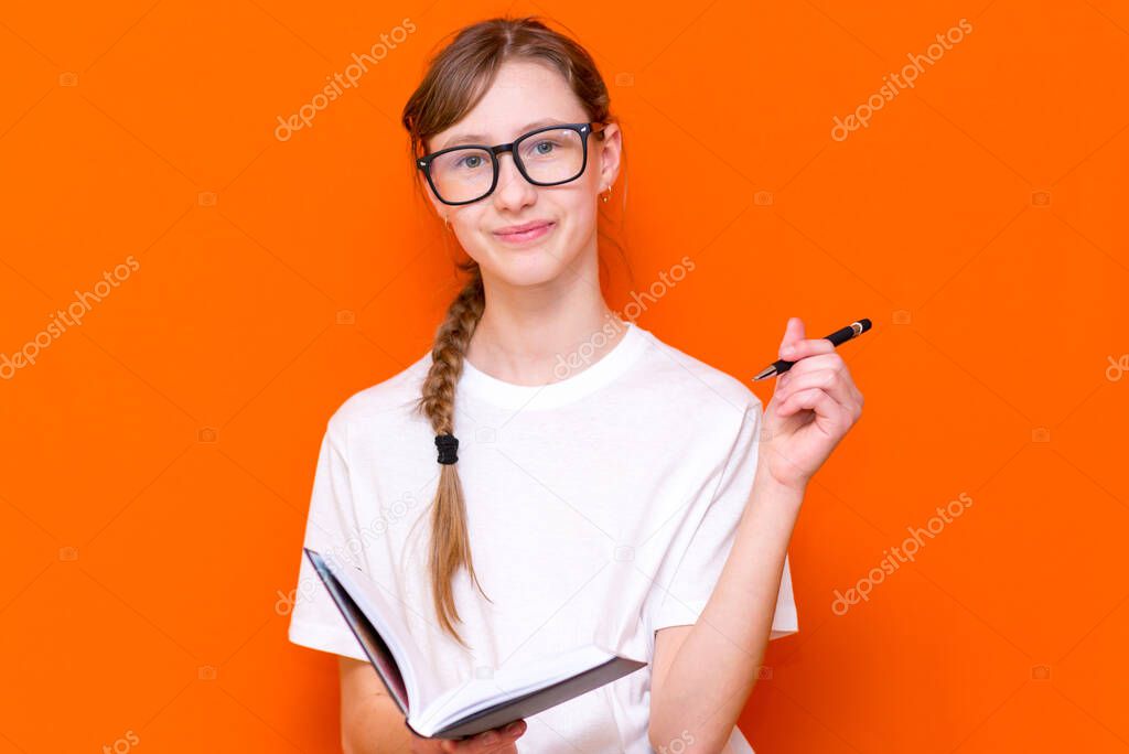 Excited thinking looking at camera young girl in eyeglasses, white T-shirt and pen something think and looking to a notebook on a orange studio background.Space for your text,banner,advertisement.