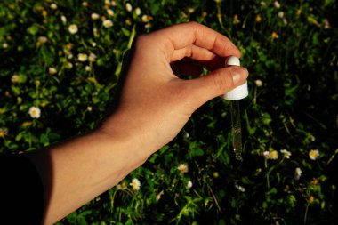Kadın eli damlatan cam şişeyi yağlı kozmetik pipetle tutuyor. Model-up. Çimenlerde küçük beyaz çiçekleri olan kadınlar için kozmetik ürün konteynırı