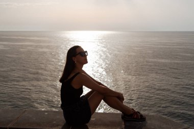 Sakin denize bakan genç bir kadın. Deniz manzarasından zevk alan genç bir kadın. Denizin üzerinde oturan mutlu ve çarpıcı bir kadının moda görüntüsü..