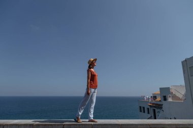 Arka manzara: uzun siyah saçlı, çarpıcı bir kız taş bir terasta duruyor ve deniz manzarasına hayran. Tropikal tatil. Dinlenme ve keyif.