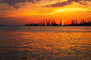 Ateşli günbatımı gökyüzü ve deniz ile güzel bir manzara. Gün doğarken kıyıda liman. Turna siluetleri ateşli, turuncu ve kırmızı gökyüzüne karşı. 