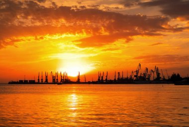 Ateşli günbatımı gökyüzü ve deniz ile güzel bir manzara. Gün doğarken kıyıda liman. Turna siluetleri ateşli, turuncu ve kırmızı gökyüzüne karşı. 