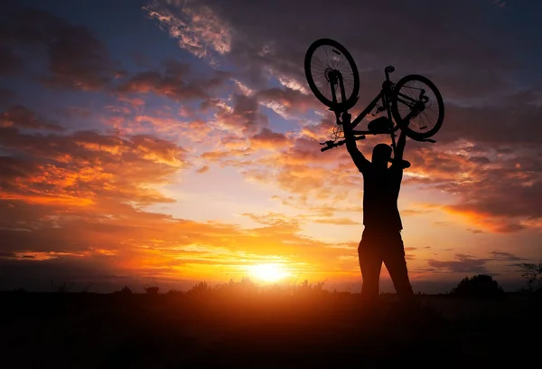 Silhueta o homem ficar em ação levantando bicicleta acima de sua hea — Fotografia de Stock
