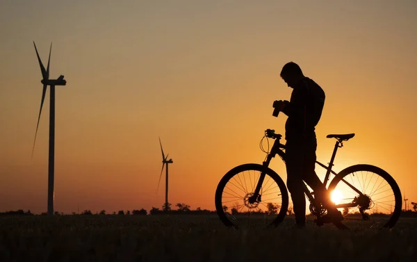 Um ciclista com uma câmera fica em um campo durante o pôr do sol agai — Fotografia de Stock