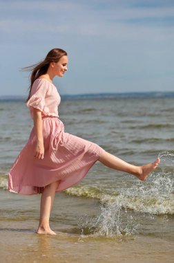 Pembe elbiseli mutlu kadın gölün kıyısında yürüyor ve suyu ayaklarıyla püskürtüyor. Neşeli bir kız denizin sahilinde durur ve ayağıyla su püskürtür..