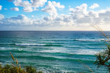 Pasifik Okyanusu'nun çarpıcı manzarası ve büyük dalgaların yuvarlanmasını bekleyen sörfçüler, mavi gökyüzü arka planı yla, Gold Coast, Avustralya'daki Burleigh Heads'ten görülebilen.