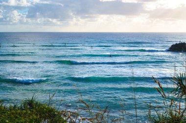 Pasifik Okyanusu'nun çarpıcı manzarası ve büyük dalgaların yuvarlanmasını bekleyen sörfçüler, mavi gökyüzü arka planı yla, Gold Coast, Avustralya'daki Burleigh Heads'ten görülebilen.