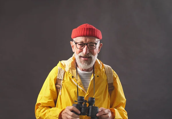 An older man with a gray beard is going on an adventure or trip with binoculars, dressed in a raincoat, raincoat, isolated background