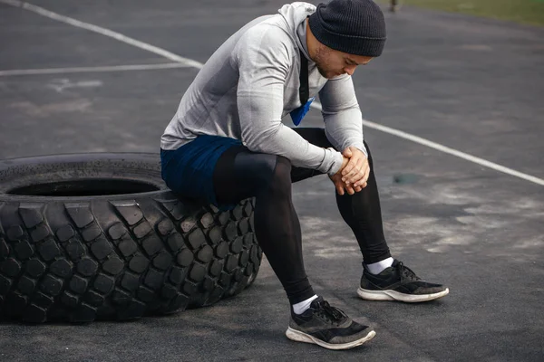 Ein Sportler ruht sich nach dem Training auf einem Sportplatz auf einem Reifen aus. Sportlicher Lebensstil. — Stockfoto