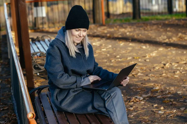 Side view of a happy girl in a coat sitting on a Park bench and using a laptop. The concept of communication.