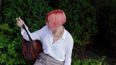 Pembe saçlı bir öğrenci korkuluğa sırt çantası koyar ve cebinden bir cep telefonu çıkarır.