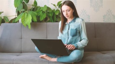 Dizüstü bilgisayarda yazan genç bir kadın evdeki kanepede oturuyor.