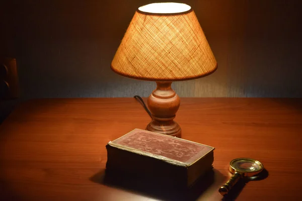 Table lamp, old book and magnifier on the desk. Soft light, reading lamp. Interior of the room. Evening time. Selective focus.