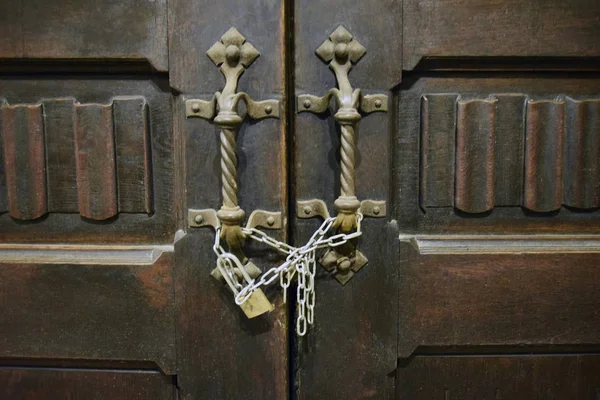 Antique wooden carved door to the temple. Bronze twisted handles entwined with an iron chain and closed on padlock. The wooden surface painted with brown paint.