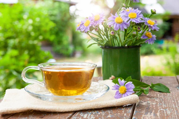 Chá de ervas úteis para a saúde na tela, em uma mesa de madeira, ao lado das flores lilás em uma caneca de metal . — Fotografia de Stock