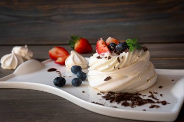 Koyu ahşap zemin üzerinde beyaz kremayla kaplı meyveli ve fındıklı bir tatlı tabağı..