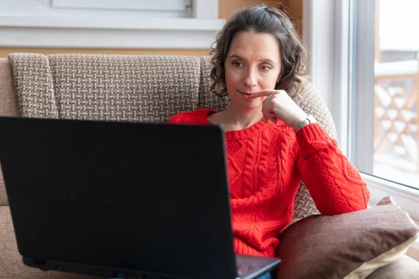 A woman in a red jacket works and watches social networks on her computer. Female with a laptop is sitting on the sofa by the window.